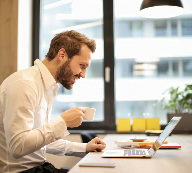 Un homme, café à la main, en train de travailler en souriant devant son ordinateur.